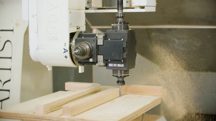 A close-up of a CNC router machine with a spindle actively cutting a piece of wood, with wood shavings flying in the background.